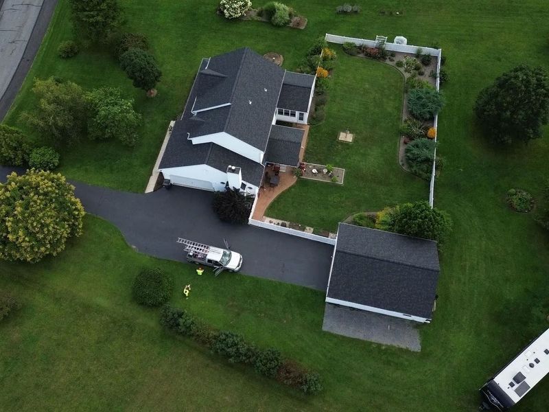 Aerial view of a house with a black roof, driveway, and surrounding green lawn. A white truck is parked outside.