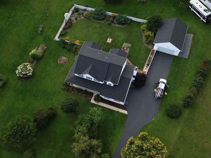 Aerial view of a house with a black roof, driveway, and detached garage on a green lawn.