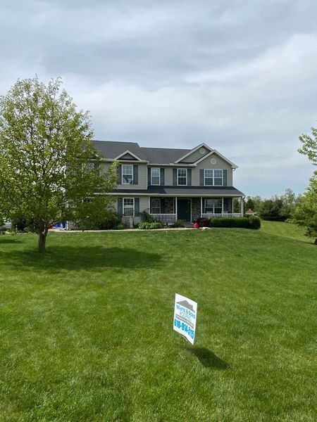 Two-story house with a light green exterior and a well-maintained lawn; a for-sale sign is in the front yard.