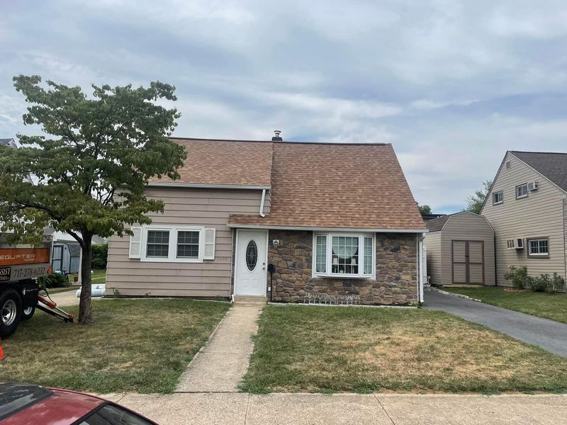 A beige house with brown roof and stone facade, tree in front, cloudy sky.