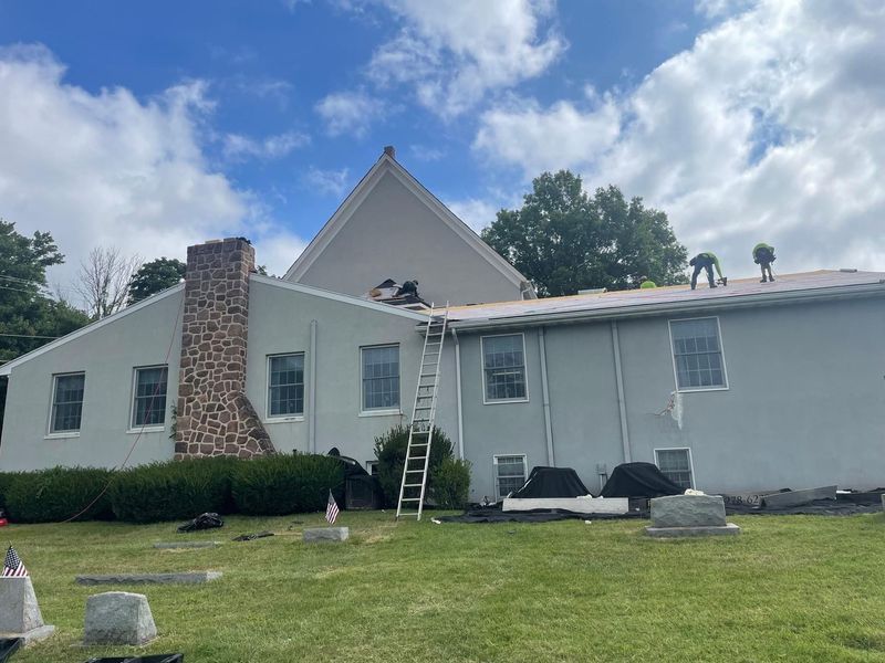 Workers on a church roof repairing shingles; gray building, blue sky, green lawn.