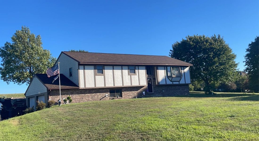 Two-story house with brown roof, white and tan facade, on grassy hill under a blue sky.