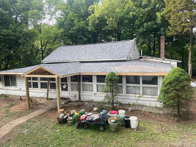 White house with gray roof, porch, and yard with junk; set in a wooded area.