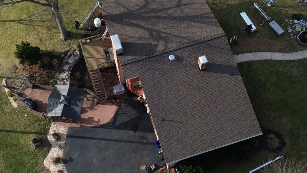 Overhead view of a brown-roofed house with a patio, on green grass; gray driveway in front.