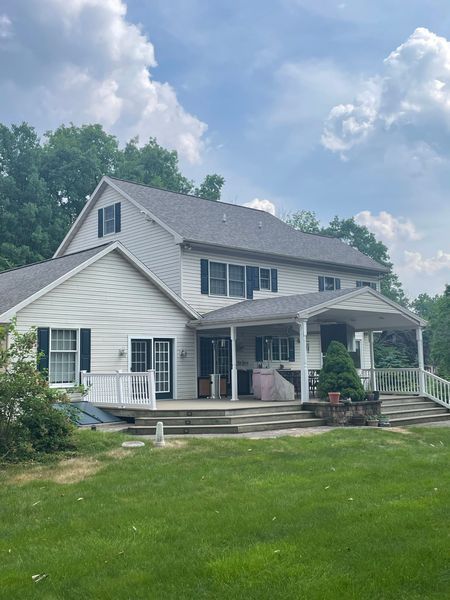 Two-story white house with a porch and gray roof, surrounded by green lawn and trees under a cloudy sky.