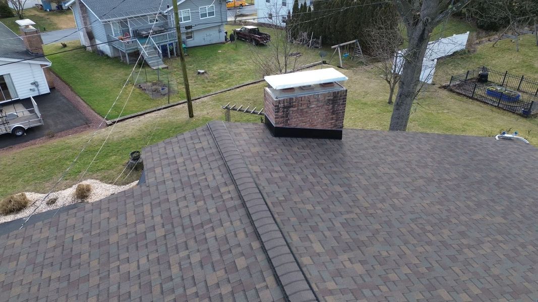 Brown shingle roof with a brick chimney. Green yard and houses in background.