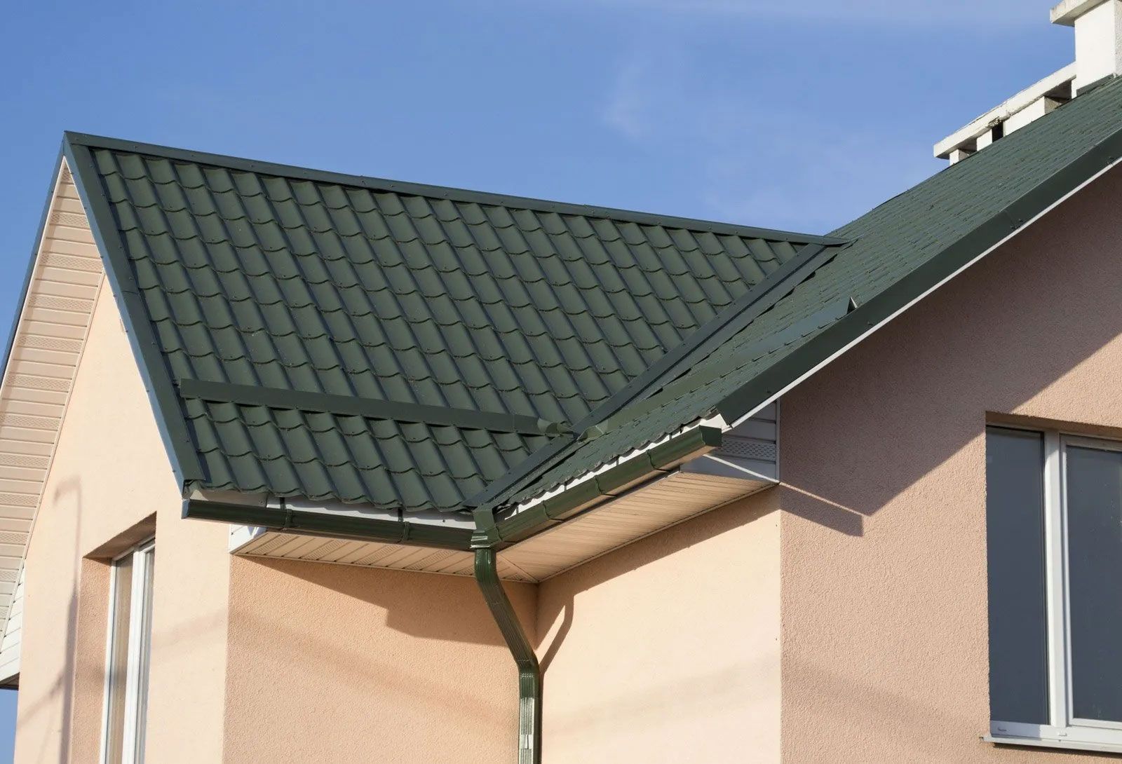 Green metal roof on a tan house with matching gutters, set against a blue sky.