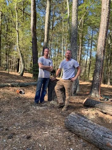 Two men in woods stand near cut logs, wearing gray shirts and jeans. One has arms crossed, the other has hand on hip.