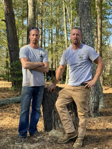 Two men standing by tree stump in a forest. One man has arms crossed, the other has a hand on his hip.