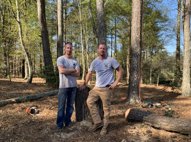 Two men in a wooded area, standing near a tree and log with a chainsaw.