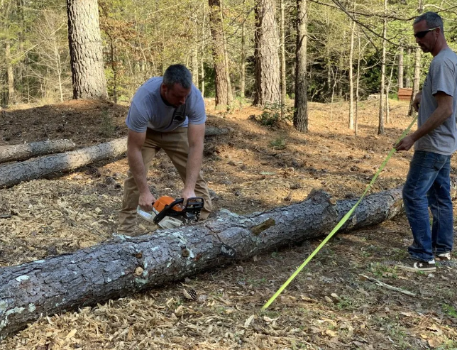 Man using a chainsaw to cut a log in a wooded area; another man watches.