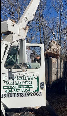 White Huckabee Tree Service truck with a person operating the lift arm and a tree stump in the bed.