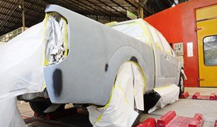 Car partially masked for paint in a repair shop, with white and yellow plastic covering and red equipment nearby