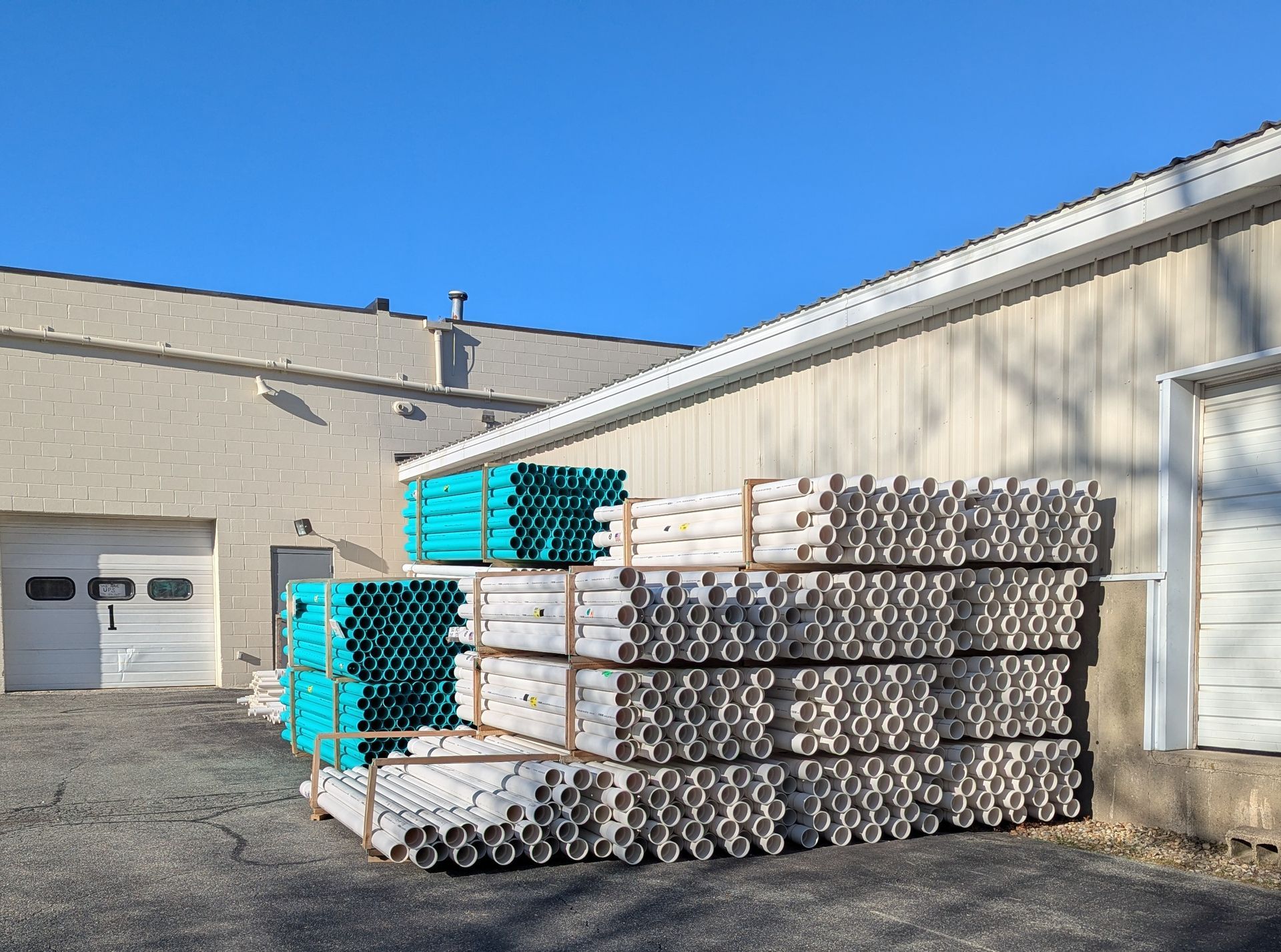 Piles of white and teal PVC pipes stacked outside a tan building with garage doors under a clear blue sky.