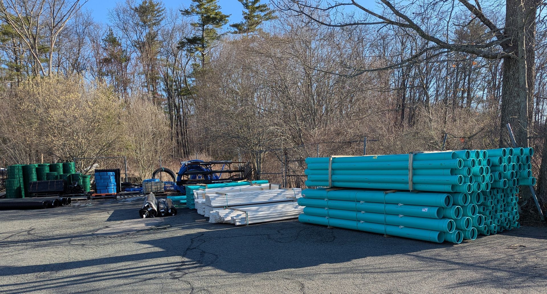 Stacks of teal pipes and other construction materials sit outdoors in a gravel yard, surrounded by trees and under a blue sky.