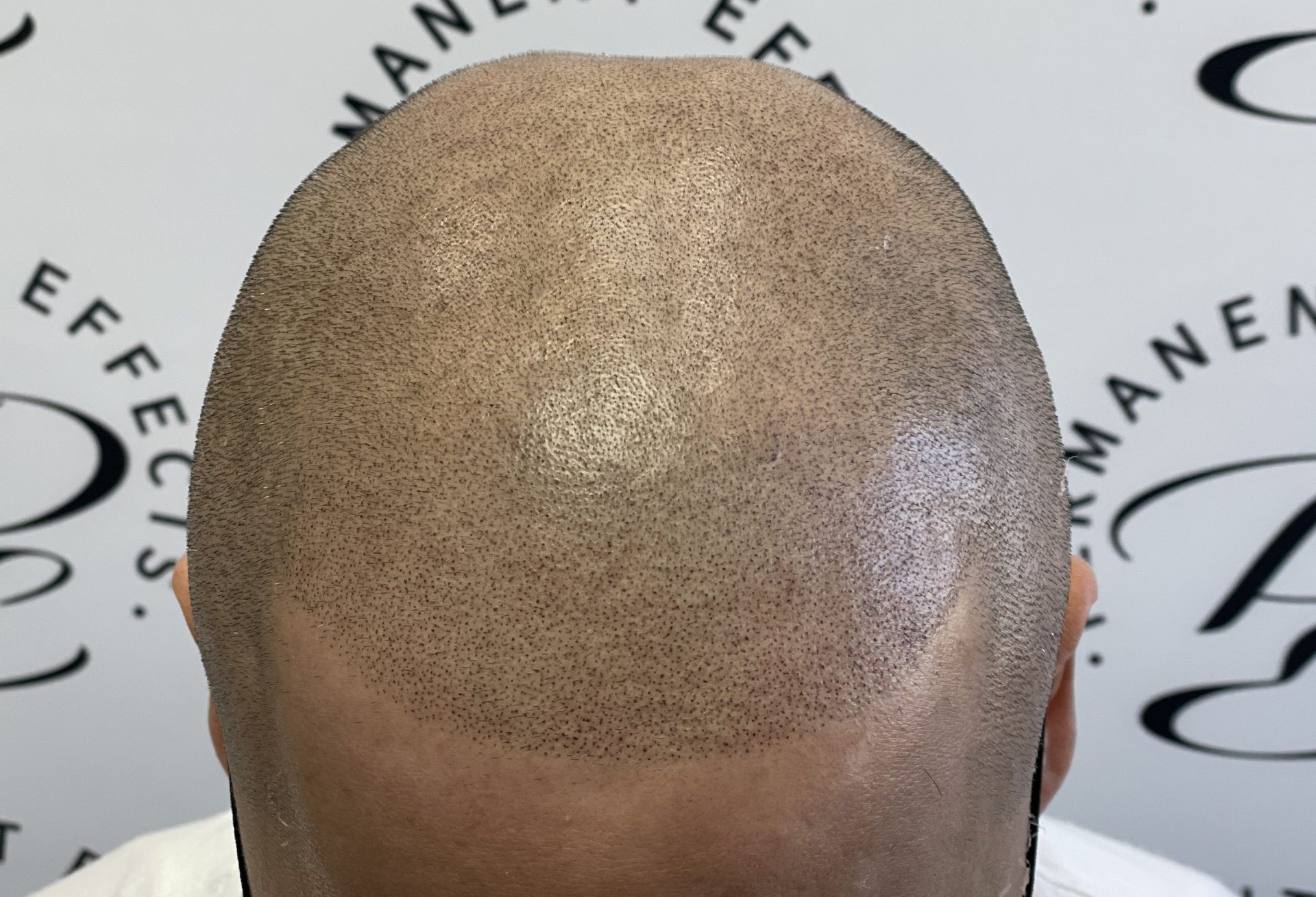 A man with a shaved head is standing in front of a wall that says ' manea ' on it