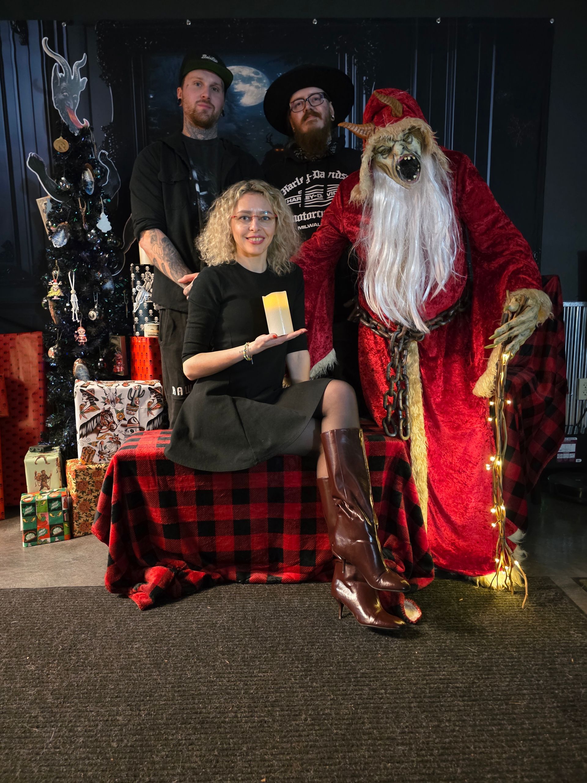 Group posing with a demonic Santa Claus; one woman holds a candle. Dark, festive setting.