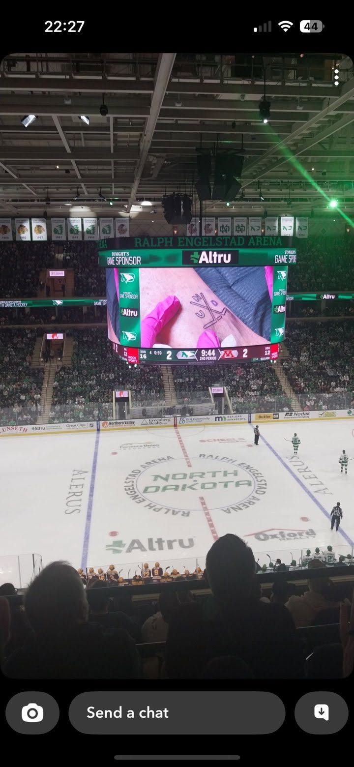 Hockey game in an arena, a tattoo is shown on the screen above the rink. Crowd watches the game.