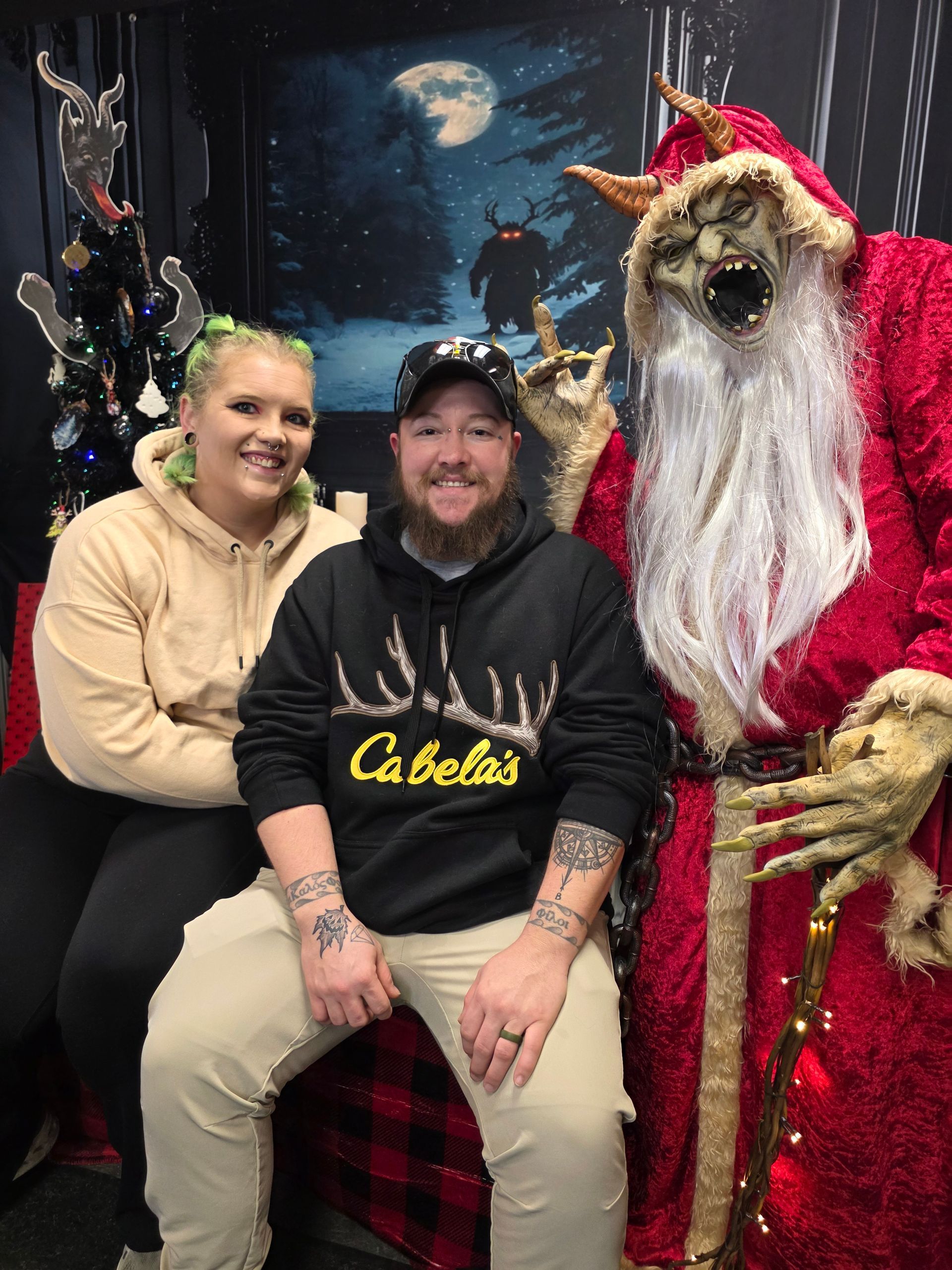 Couple poses with Krampus in a dark, festive setting. Krampus is dressed in red with horns.
