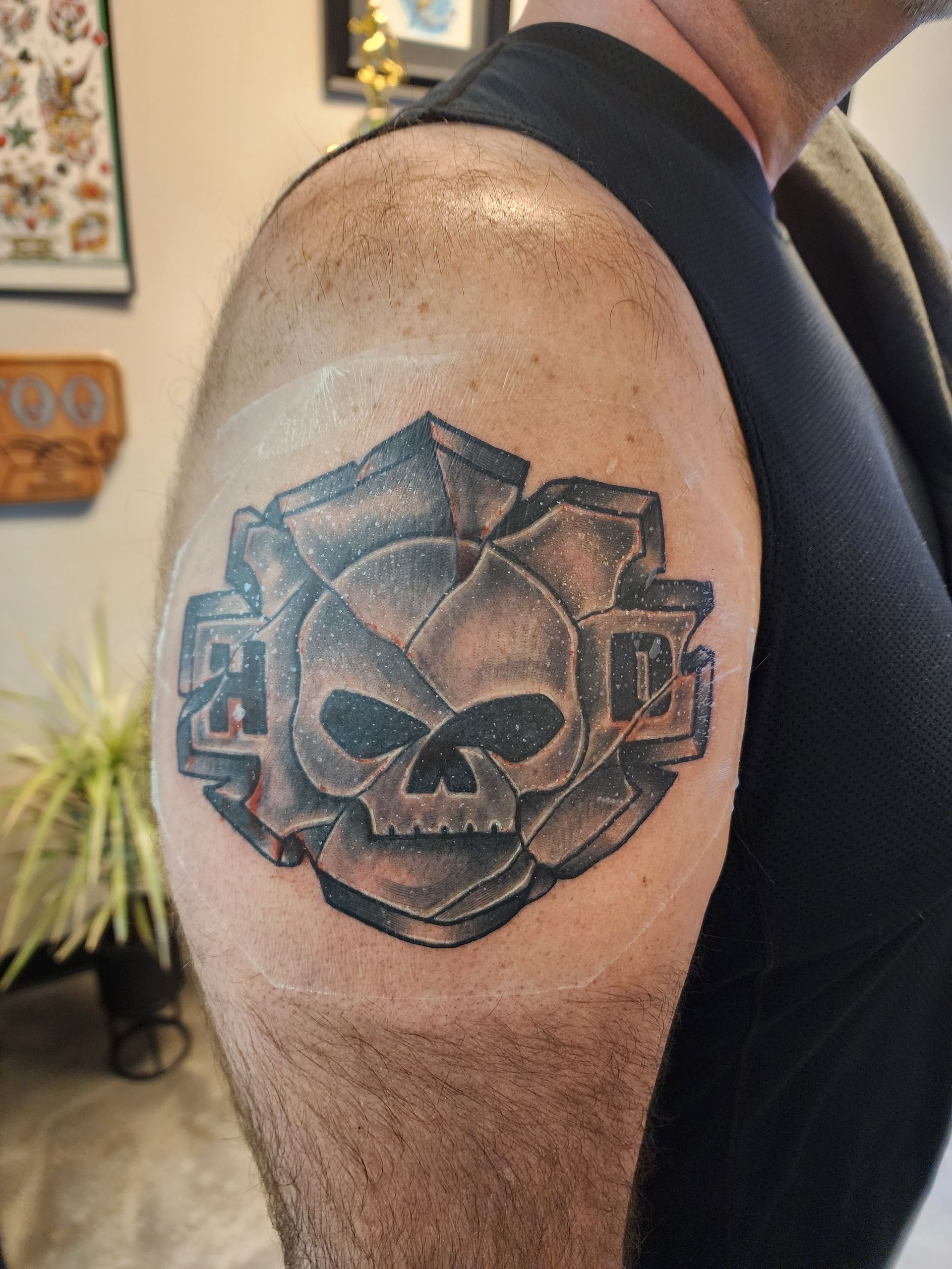 Tattoo of a skull within a stylized rose on a person's shoulder. Black tank top, tattoo parlor backdrop.