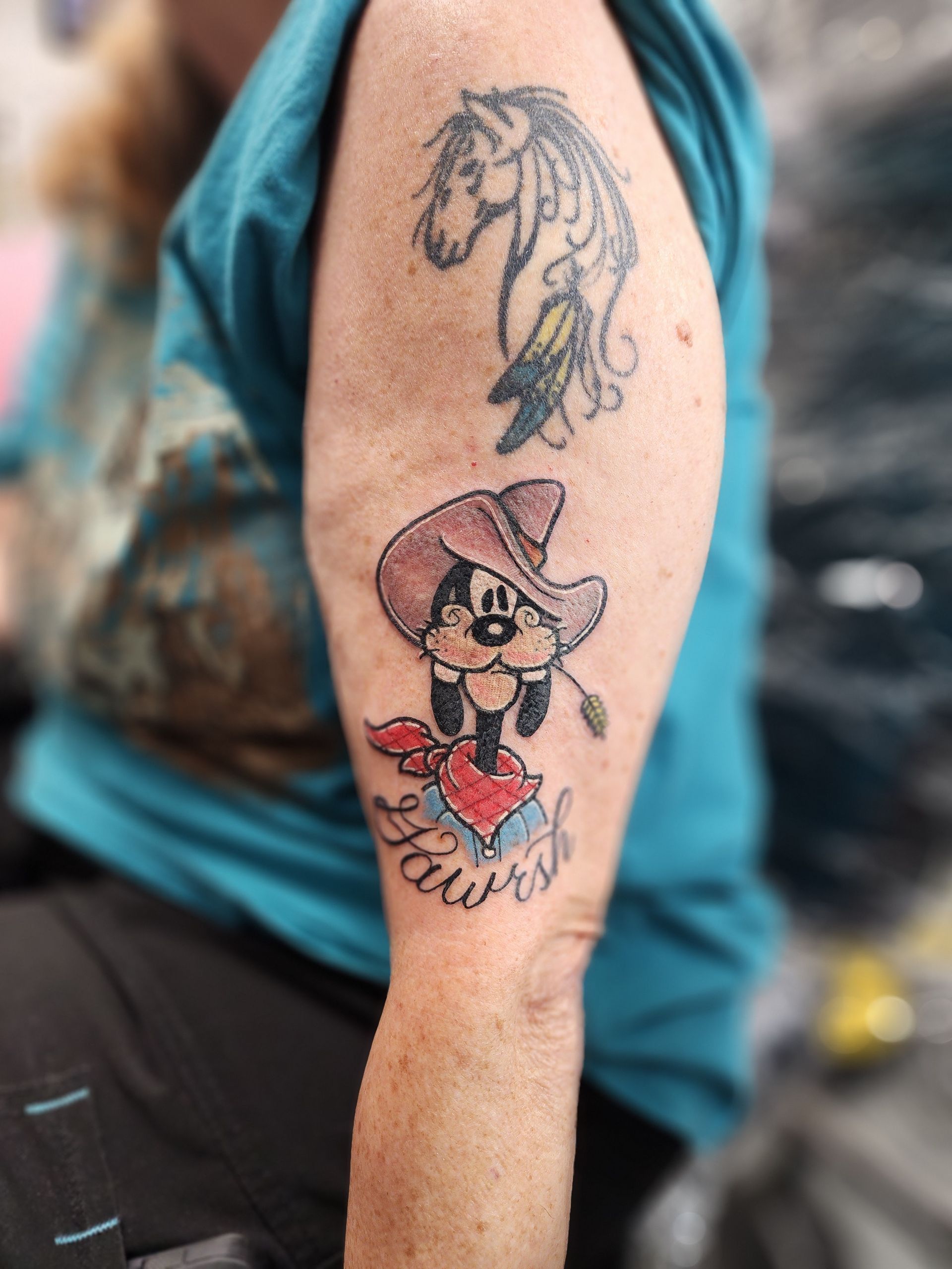 Tattoo of a horse and Goofy in a cowboy hat on a person's arm. Below Goofy is the word