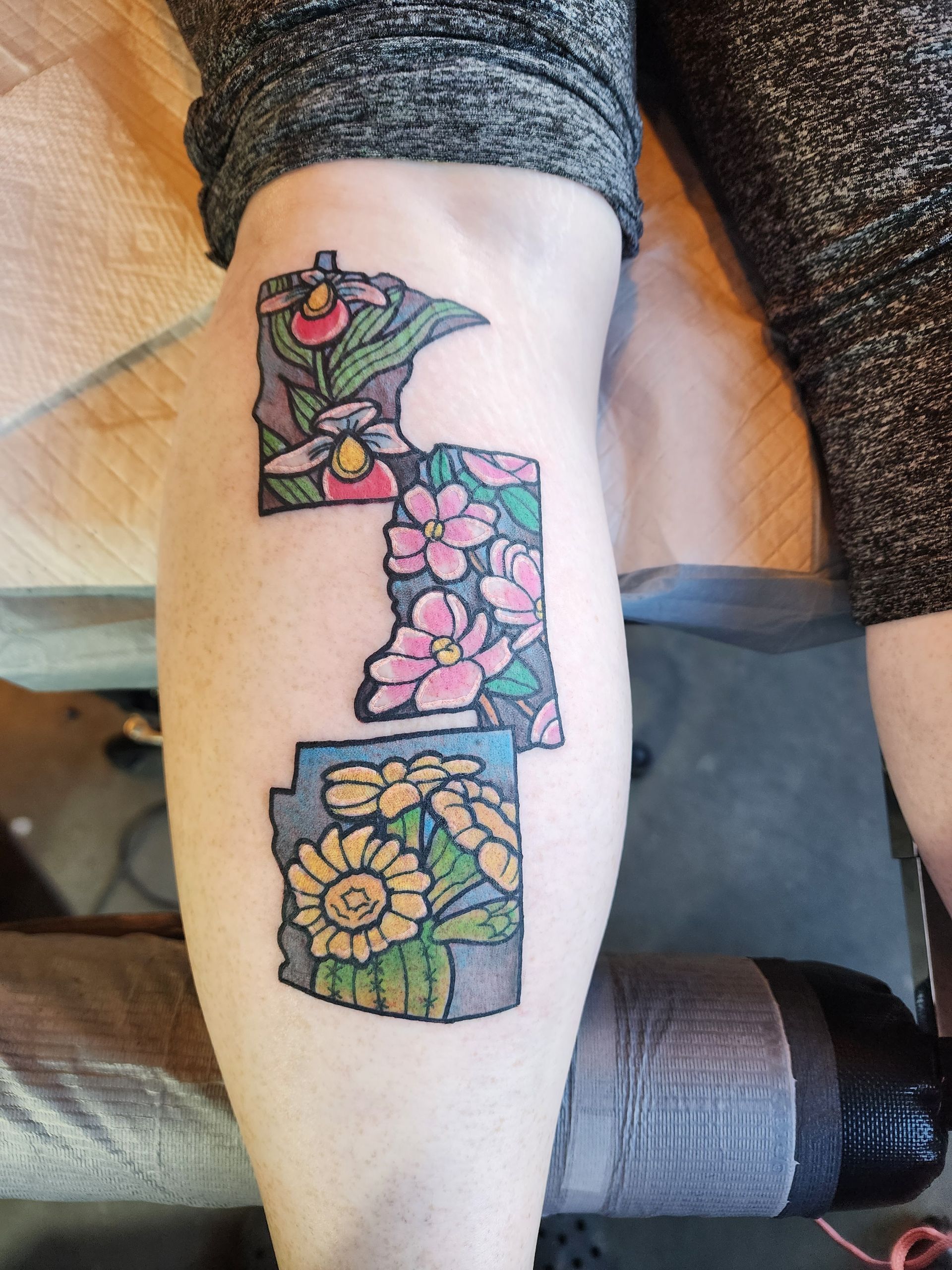 Tattoo on leg: Three US state outlines with flowers, including pink blossoms and yellow sunflowers.