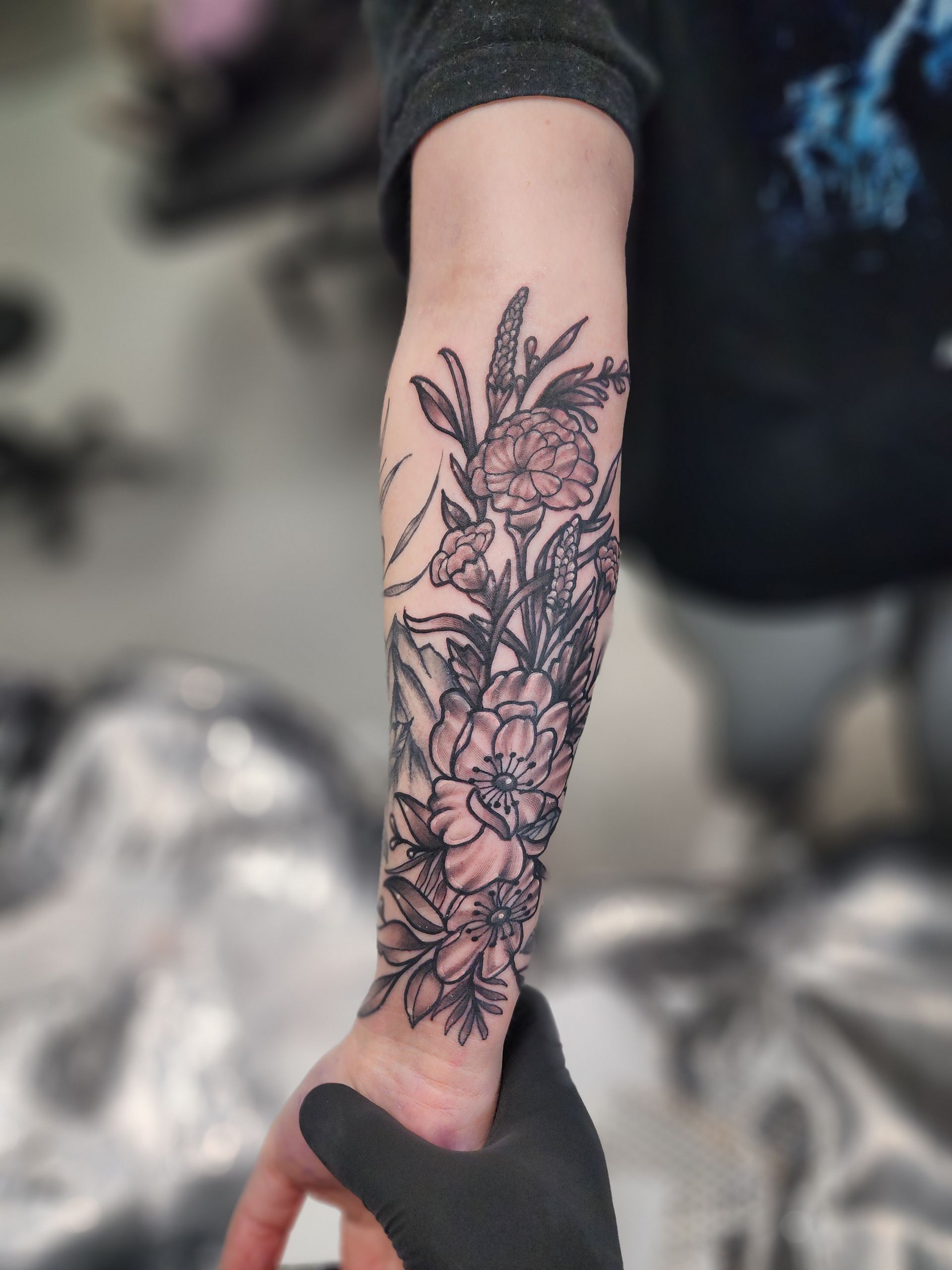 Black and white floral tattoo on a person's forearm, covering wrist to elbow. Gloved hand holds wrist.