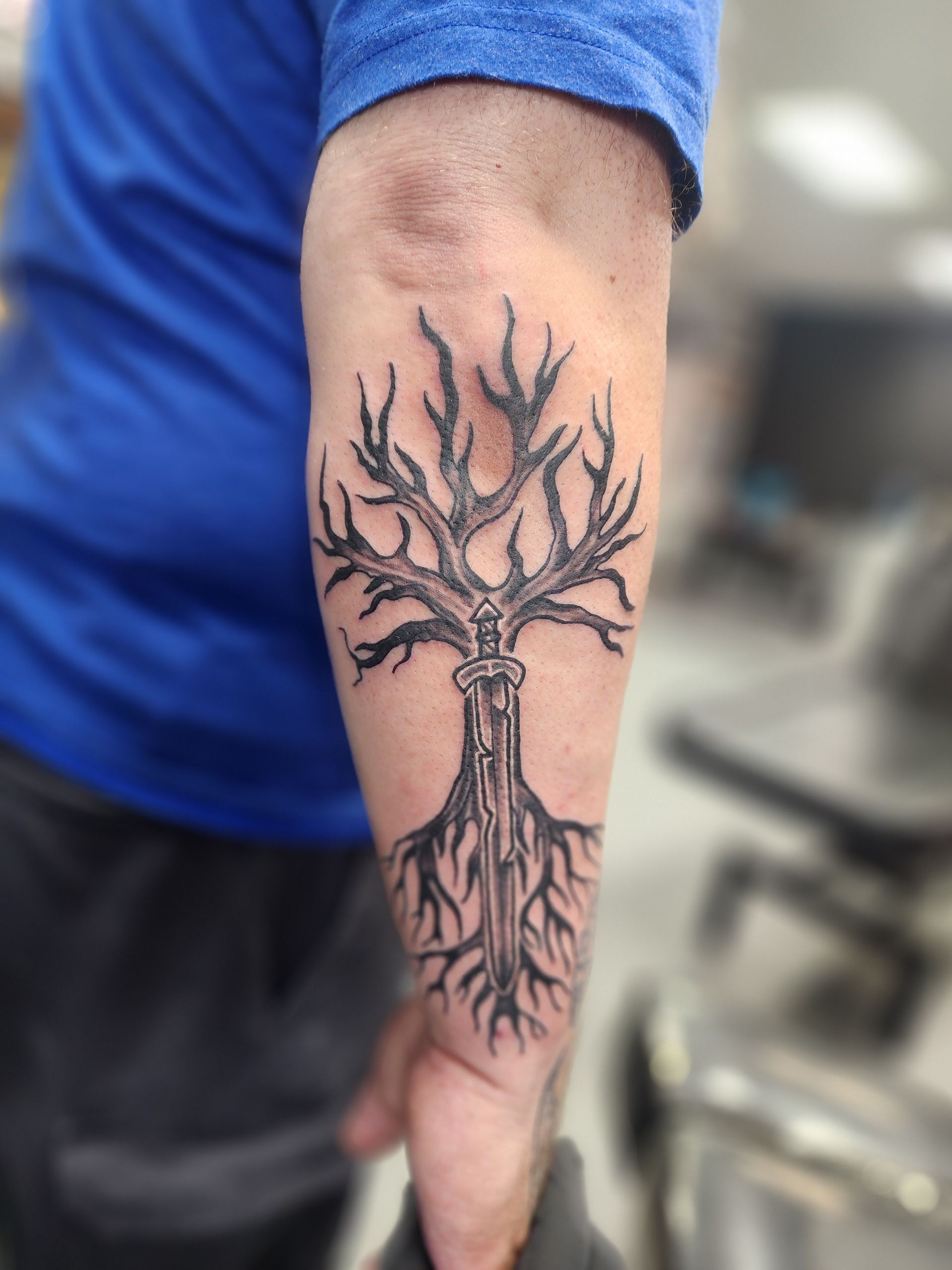 Arm tattoo of a tree with a sword in the trunk; black ink on skin.