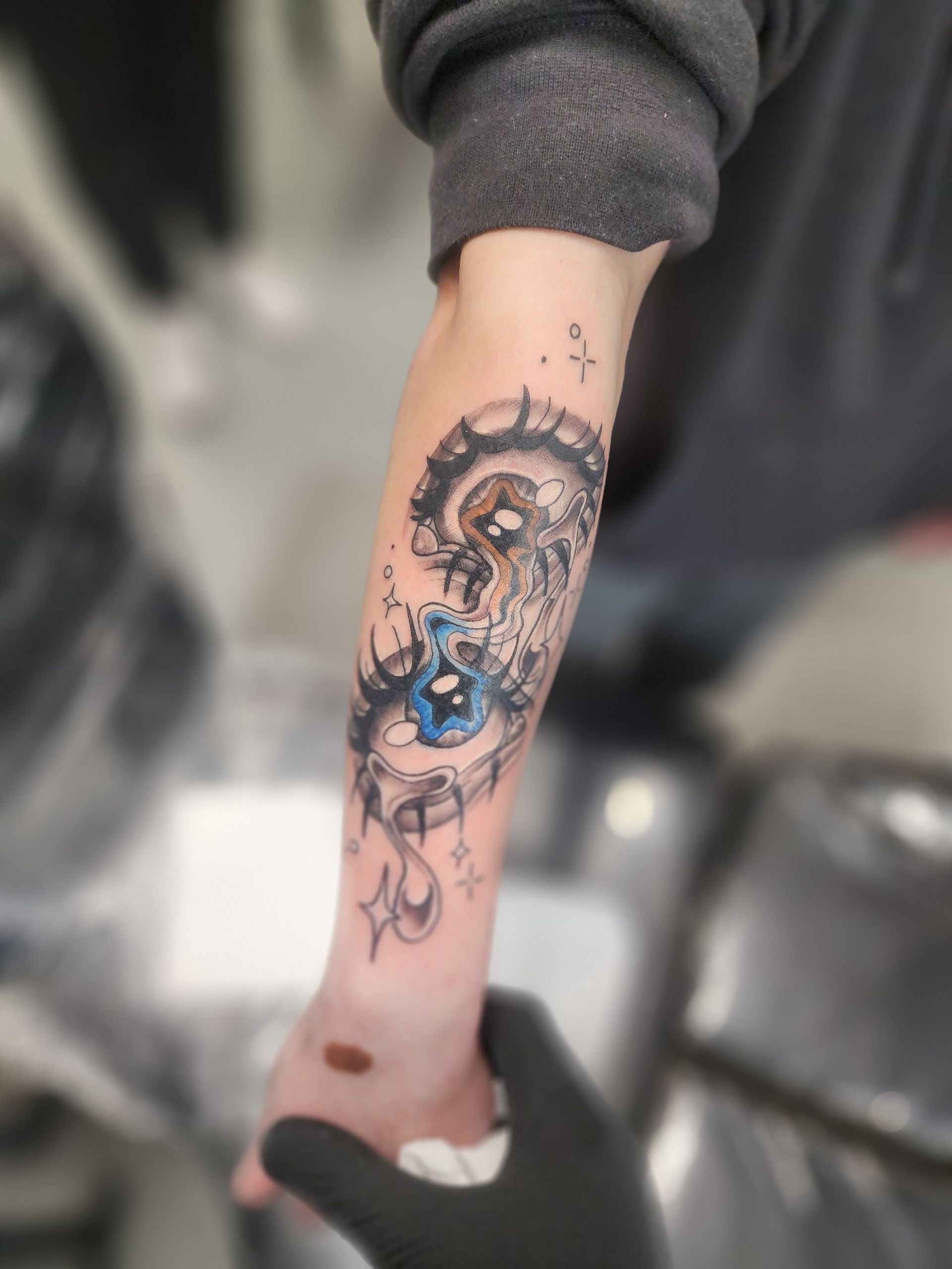 Tattoo on forearm, featuring an eye with a blue iris, surrounded by black and gray designs.