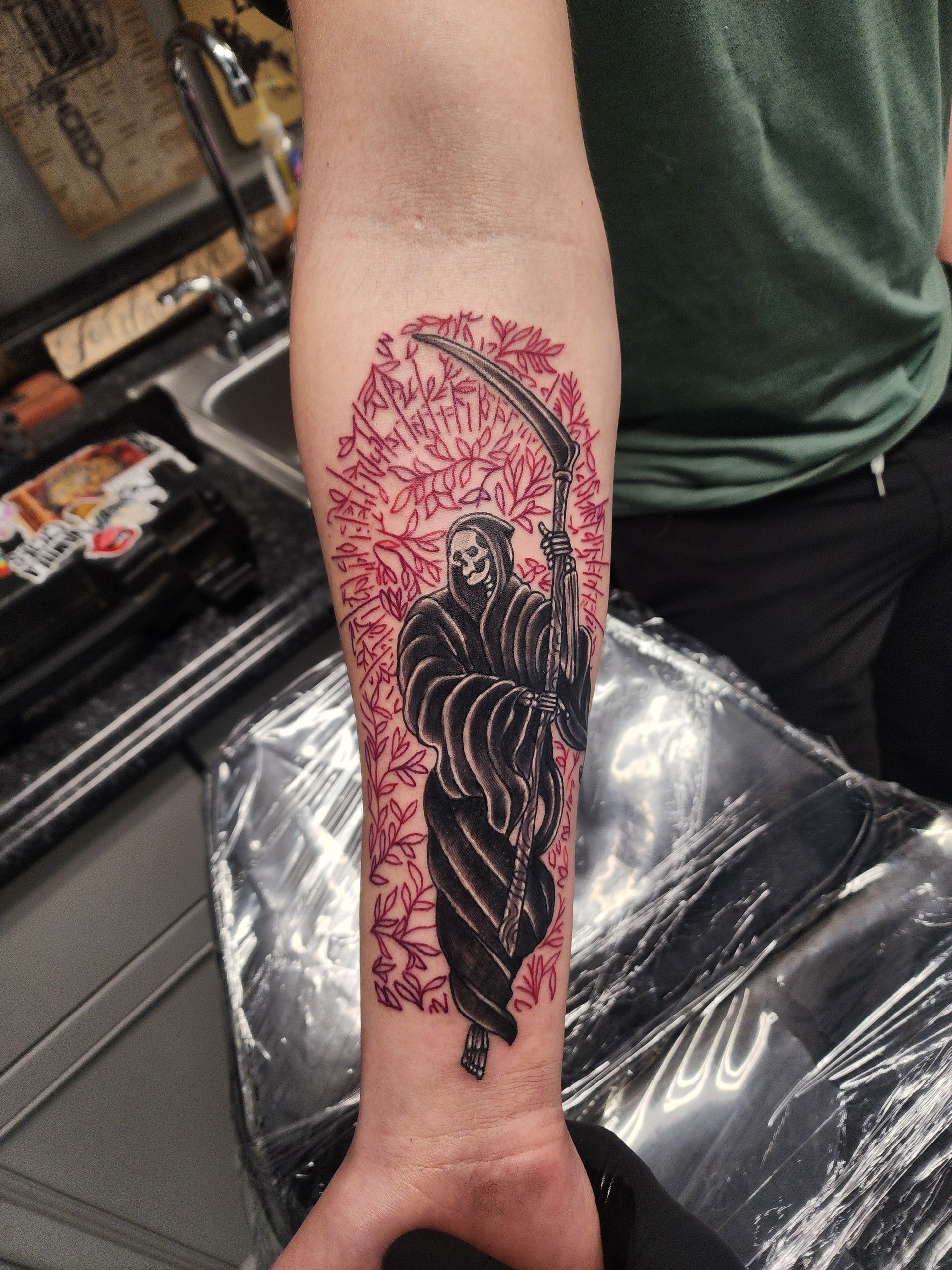 Tattoo of the grim reaper with a scythe, black silhouette on a red floral background, on an arm.