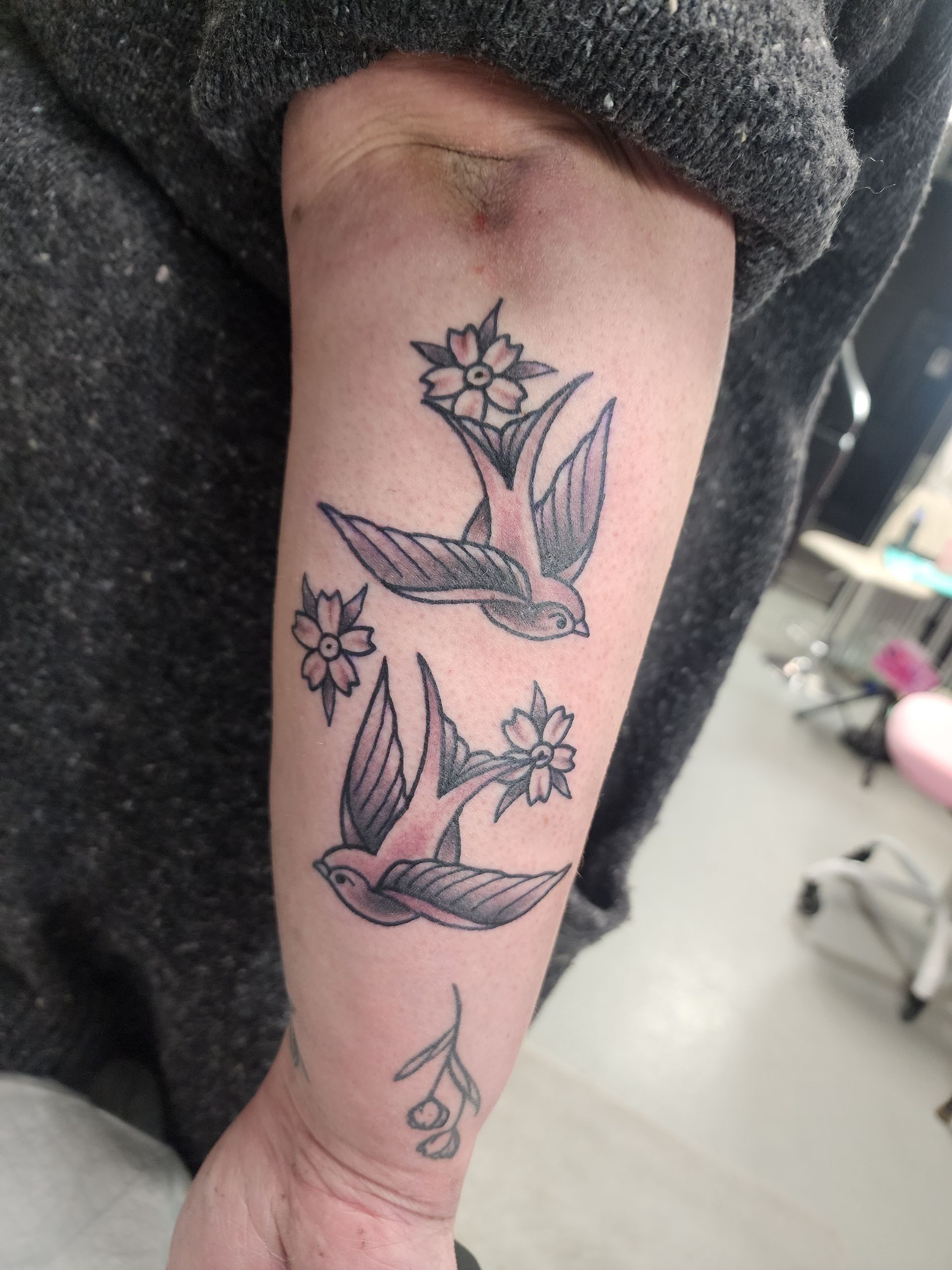 Tattoo of two swallows with flowers on forearm, with small olive branch below. Grey and purple ink.