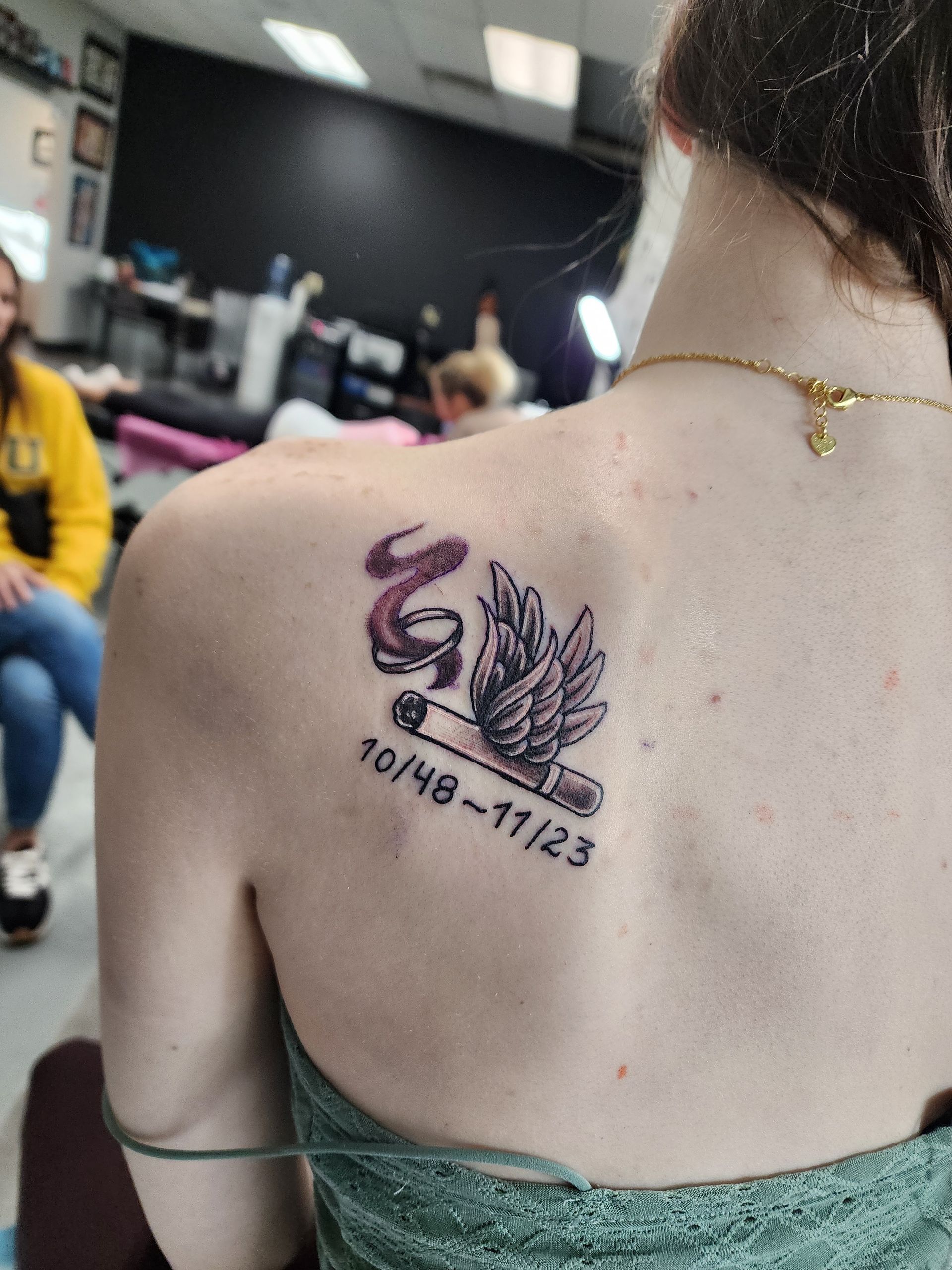 Woman's back with a memorial tattoo: winged cigarette with ribbon and dates 10/48-11/23. In a tattoo studio.