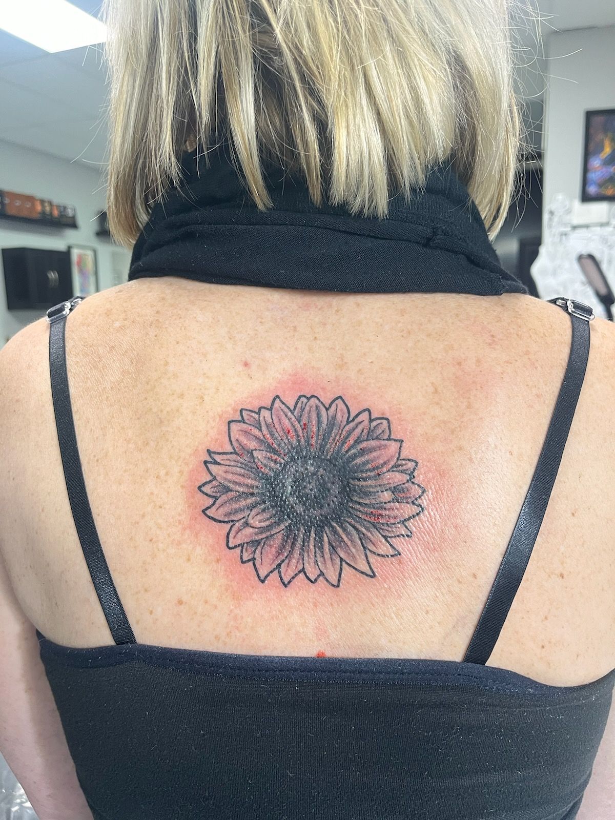Back view of a person with a black strap top, showing a sunflower tattoo on their upper back.