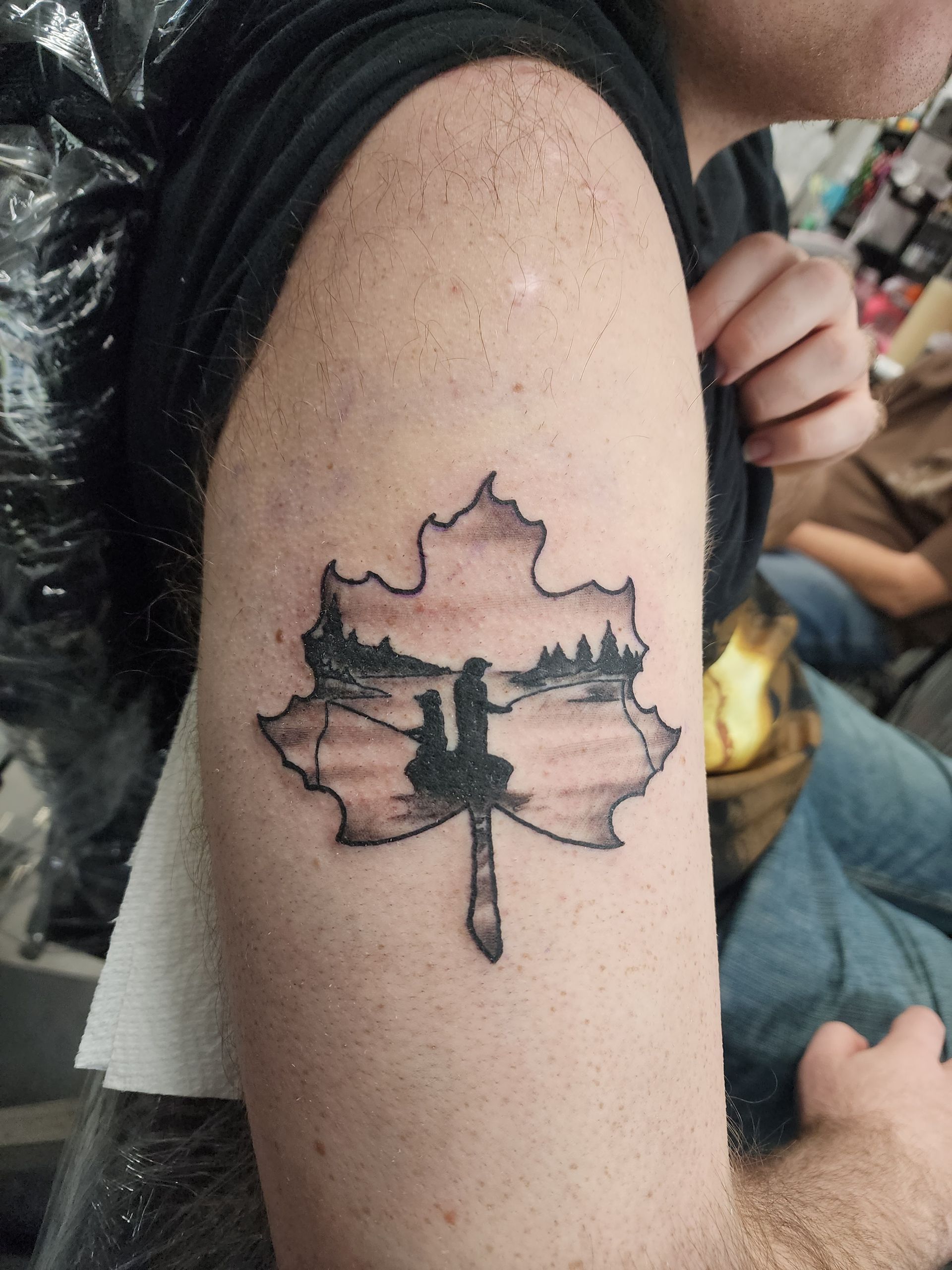 Tattoo of a landscape scene inside a maple leaf outline on a person's upper arm.