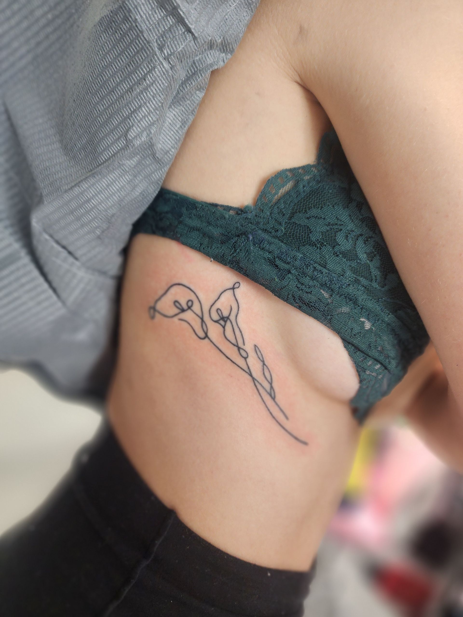 Tattoo of two calla lilies on a person's side, near a green lace bra and dark pants.