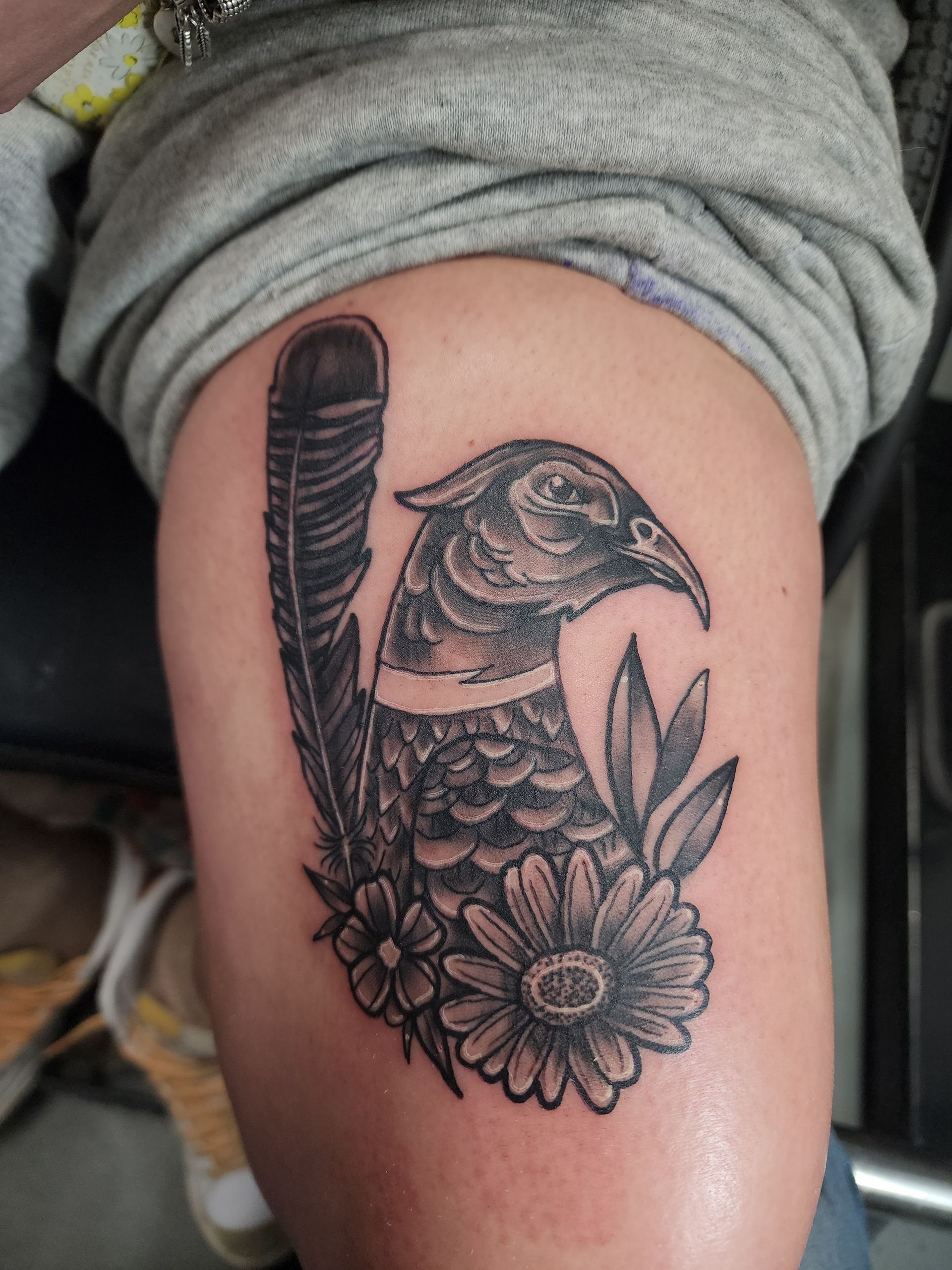 Tattoo of a pheasant with feather, flowers, and foliage on a person's thigh in black and gray.