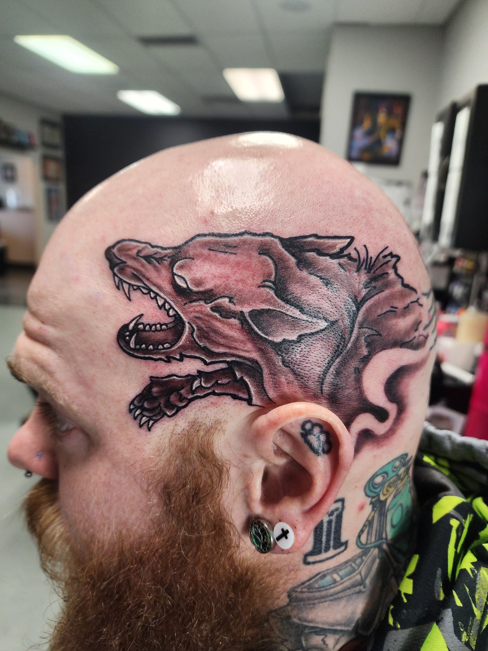 Person with a bald head, wolf tattoo on the side, and beard. Tattoo is red and gray, mouth open.