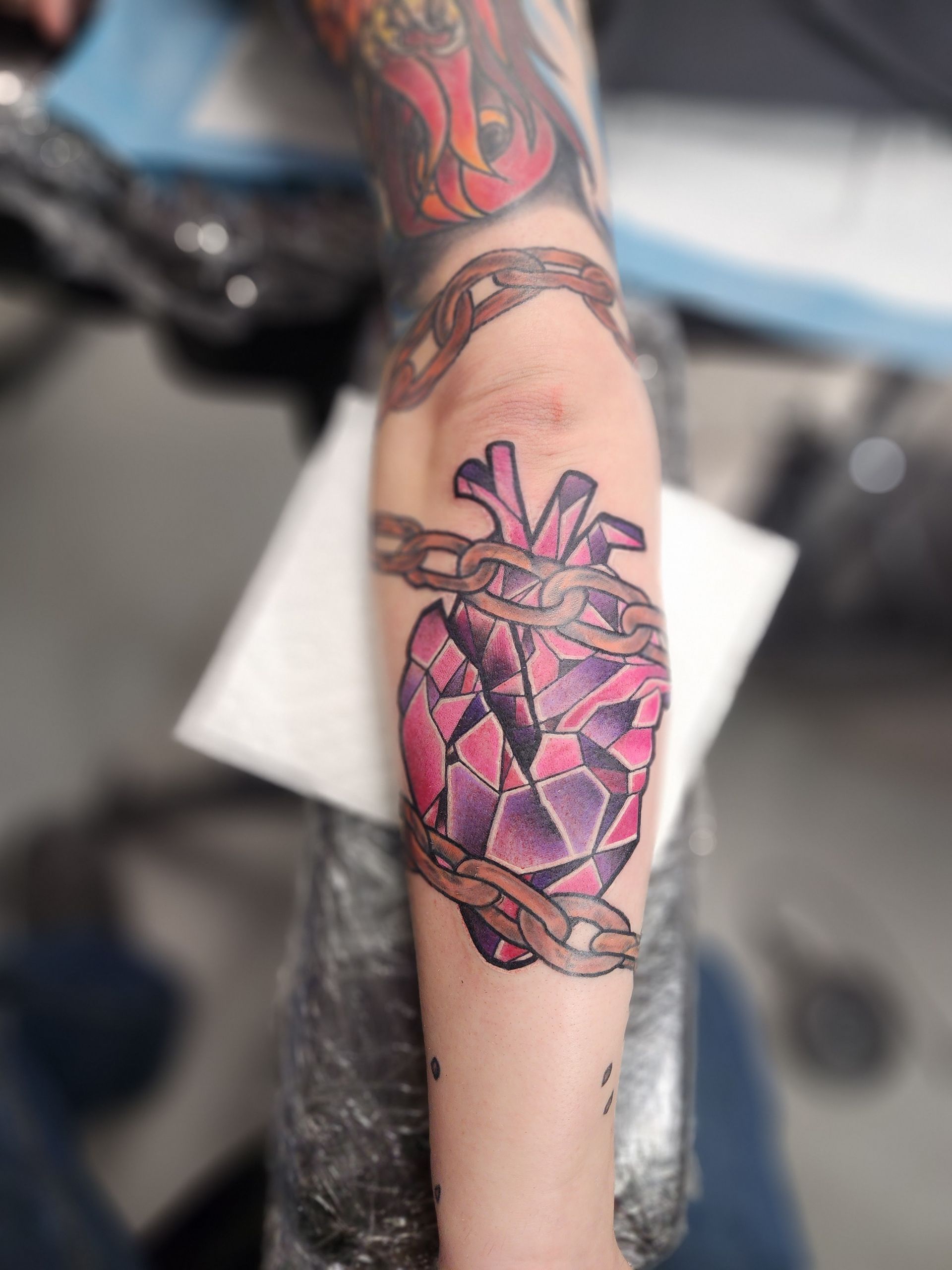 Tattoo of a broken heart, purple and pink geometric shapes, wrapped in a chain, on a person's arm.
