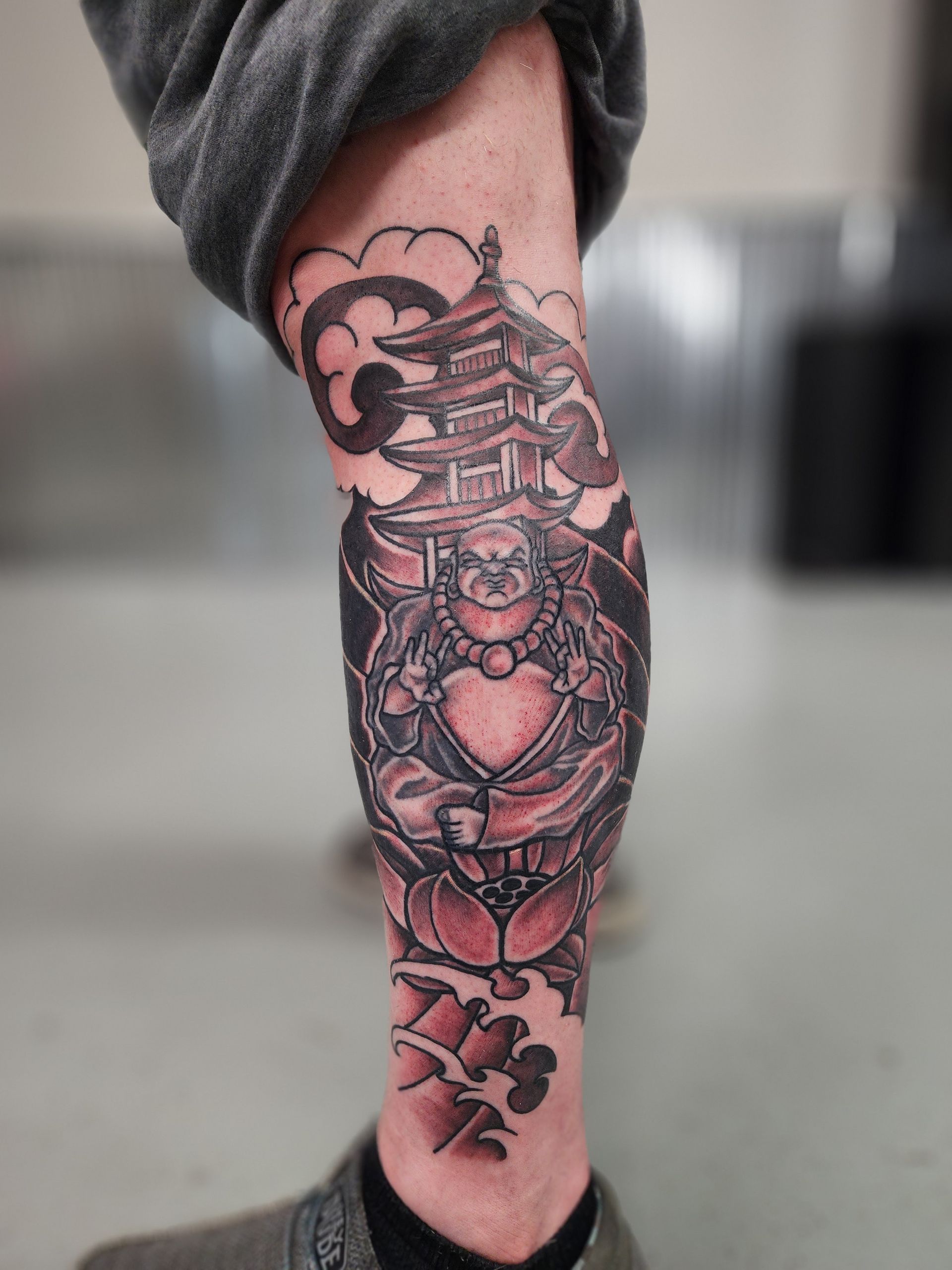 Black and gray leg tattoo of a seated Buddha figure with pagoda and clouds; red lotus at ankle.