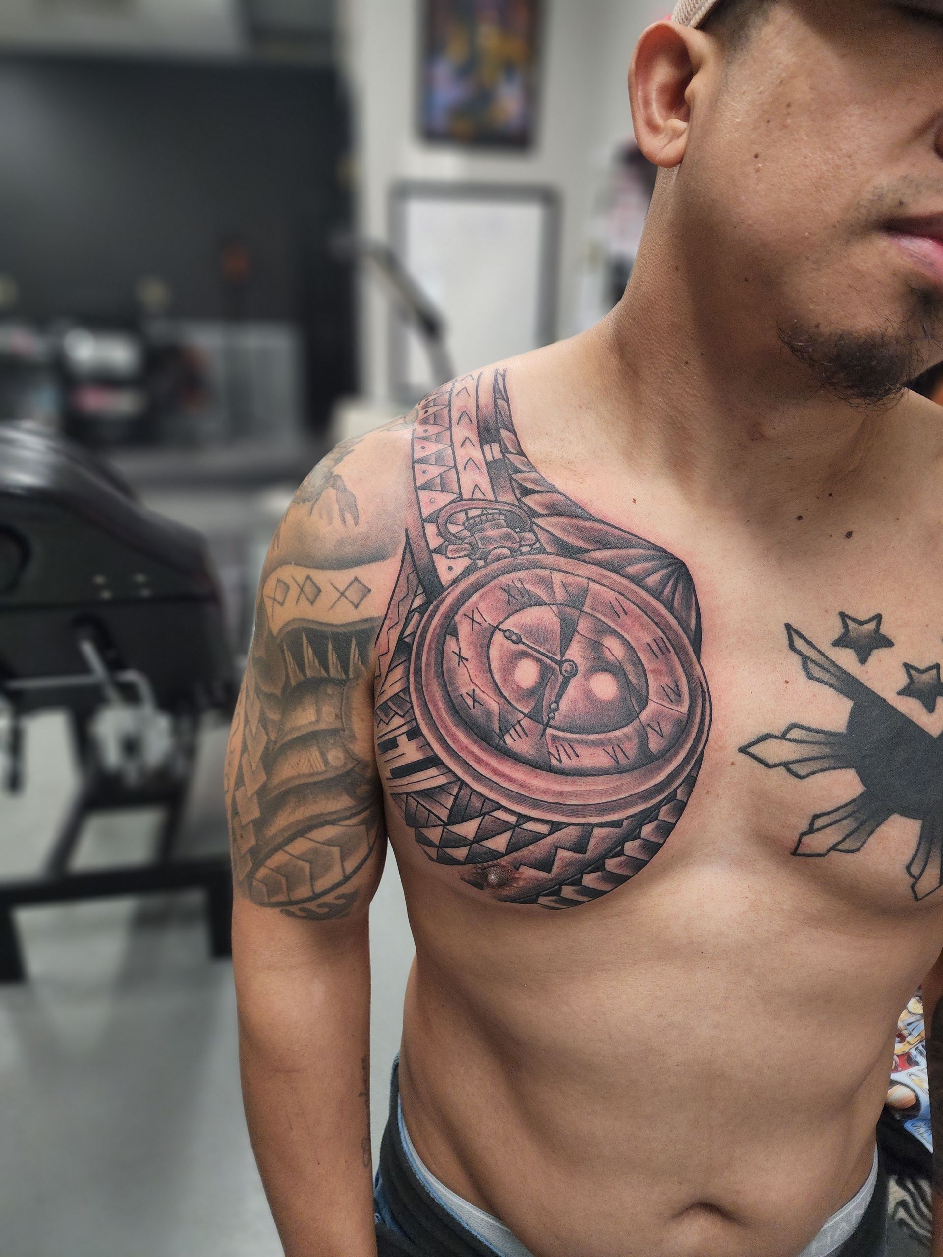 Man with chest tattoo featuring a circular design and other Polynesian-style ink work on his shoulder.