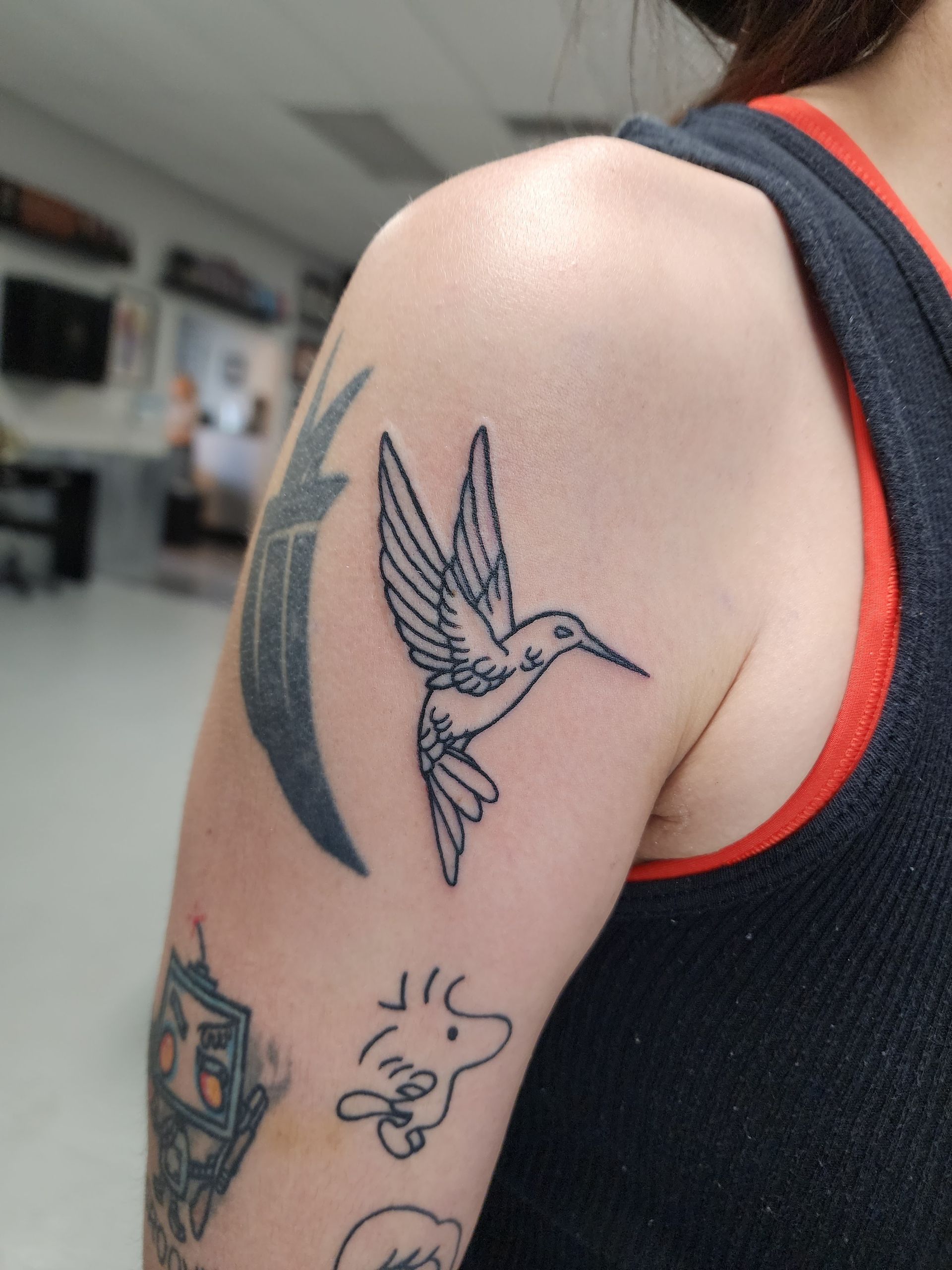 Tattoo of a hummingbird in flight on a person's upper arm. Black ink outlines on skin.