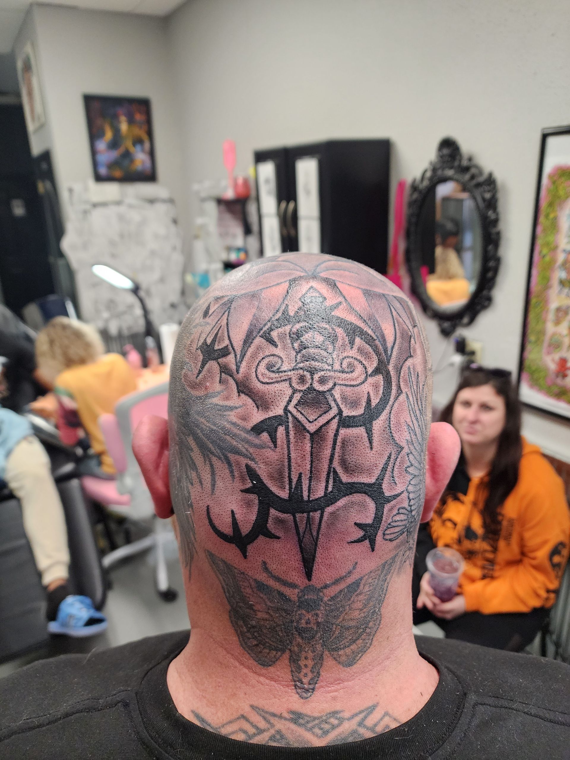 Man's head with large black tattoo. In a tattoo parlor, a person with orange shirt watches.