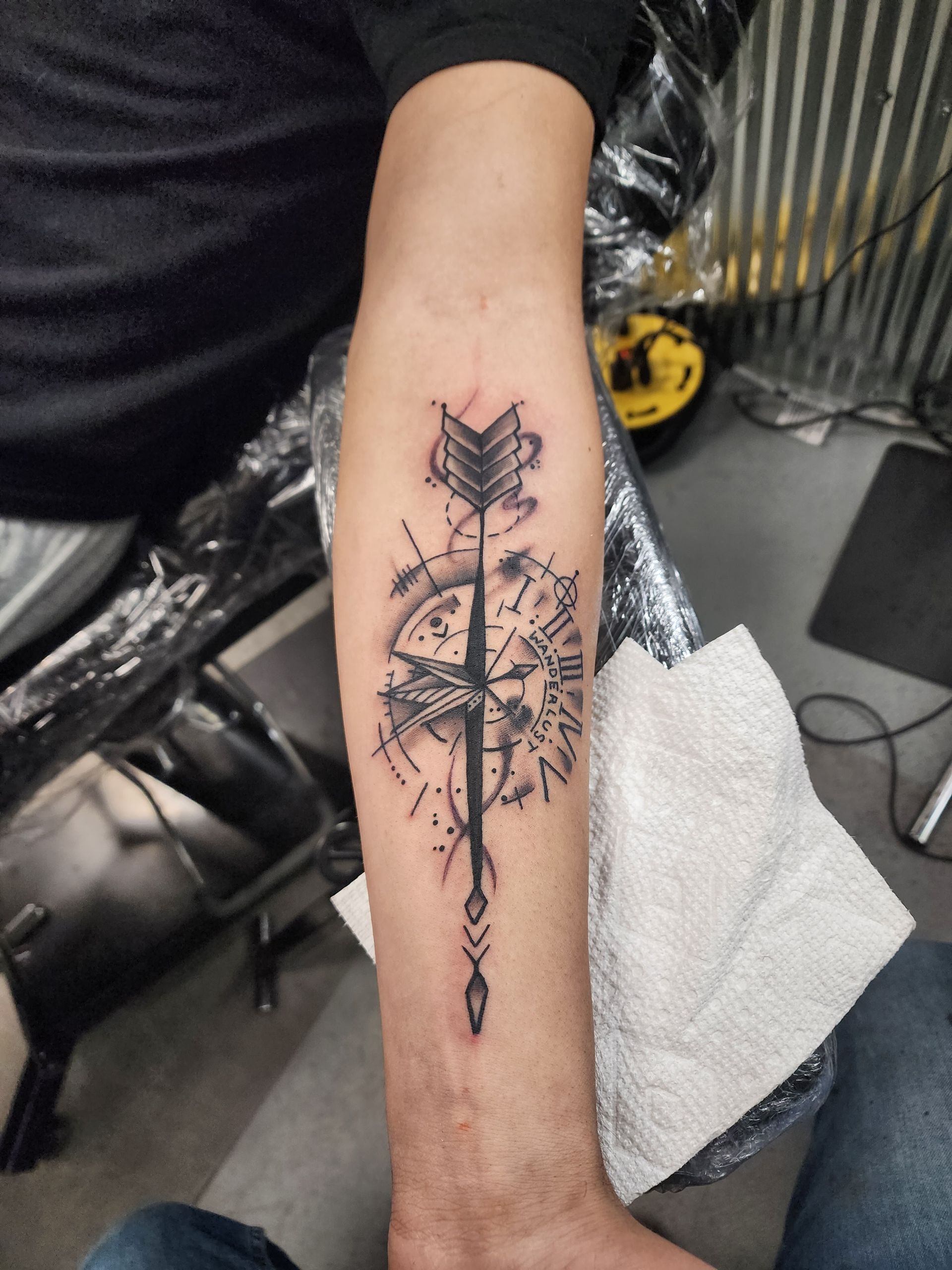 Tattoo of a compass and arrow on a person's forearm, black ink, in a tattoo parlor setting.