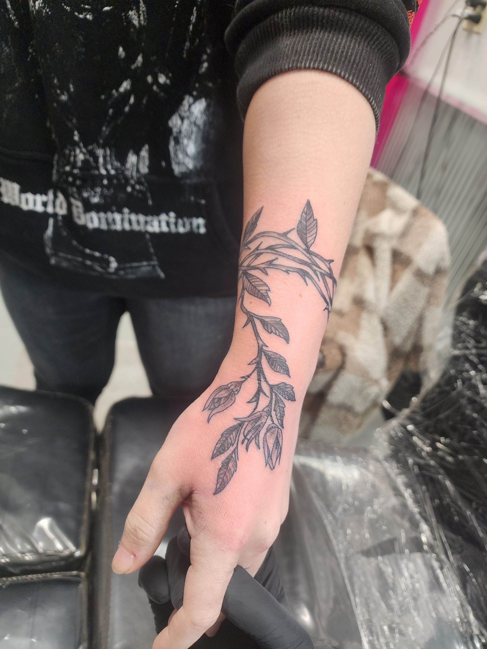 Person's arm with a tattoo of a leafy vine on the wrist and forearm, in a tattoo parlor setting.