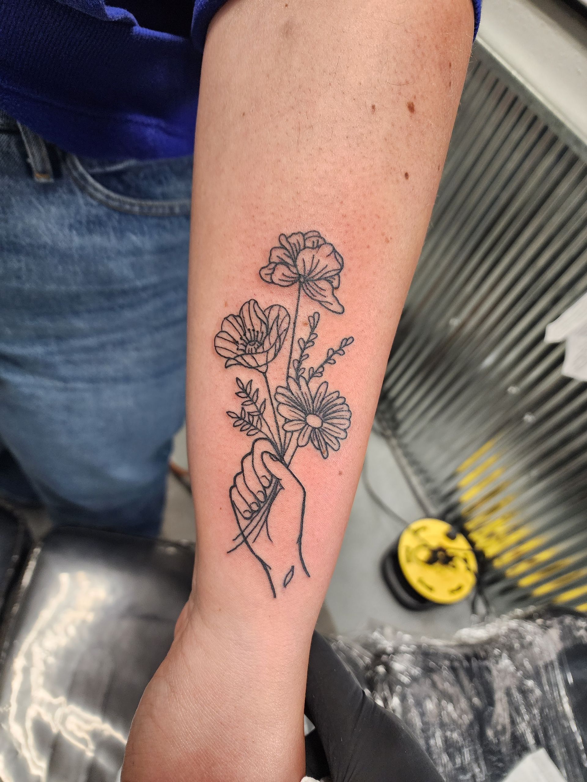 Black-ink tattoo of a hand holding a bouquet of flowers on an arm.