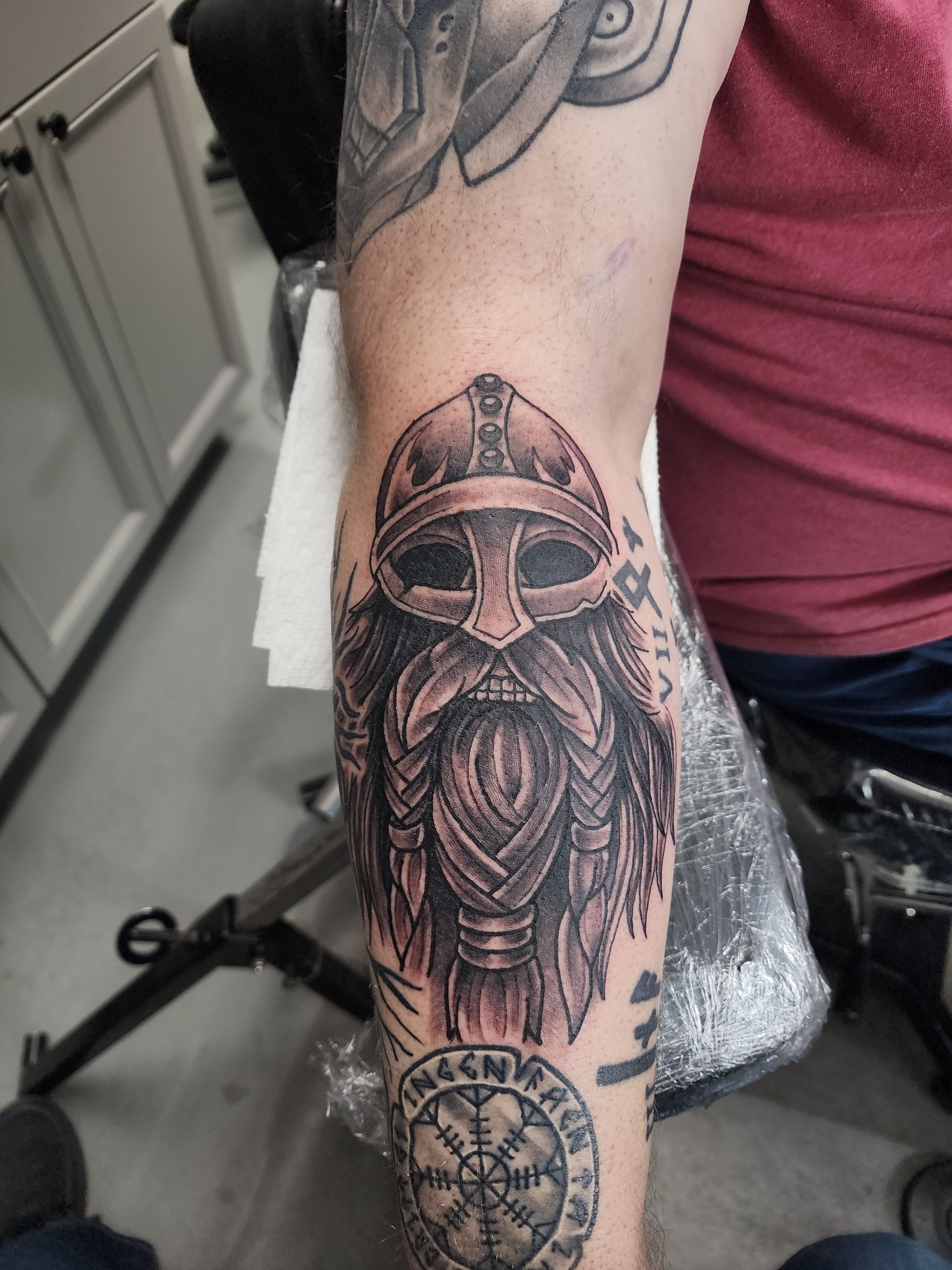 Arm tattoo: Viking helmet and braided beard in black ink, above a compass symbol.