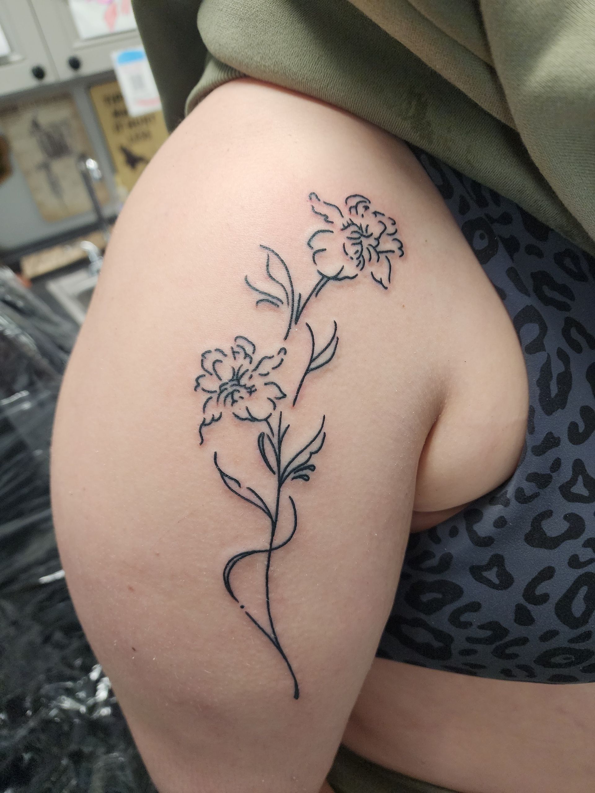 Black line art floral tattoo on a person's upper thigh, with leaves and a curving stem.