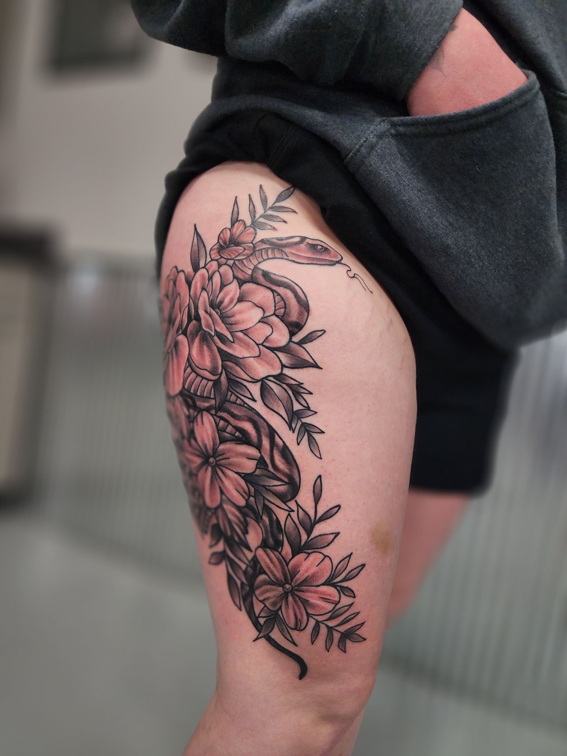 Tattoo of flowers and foliage, shaded in black and gray, on a person's thigh.