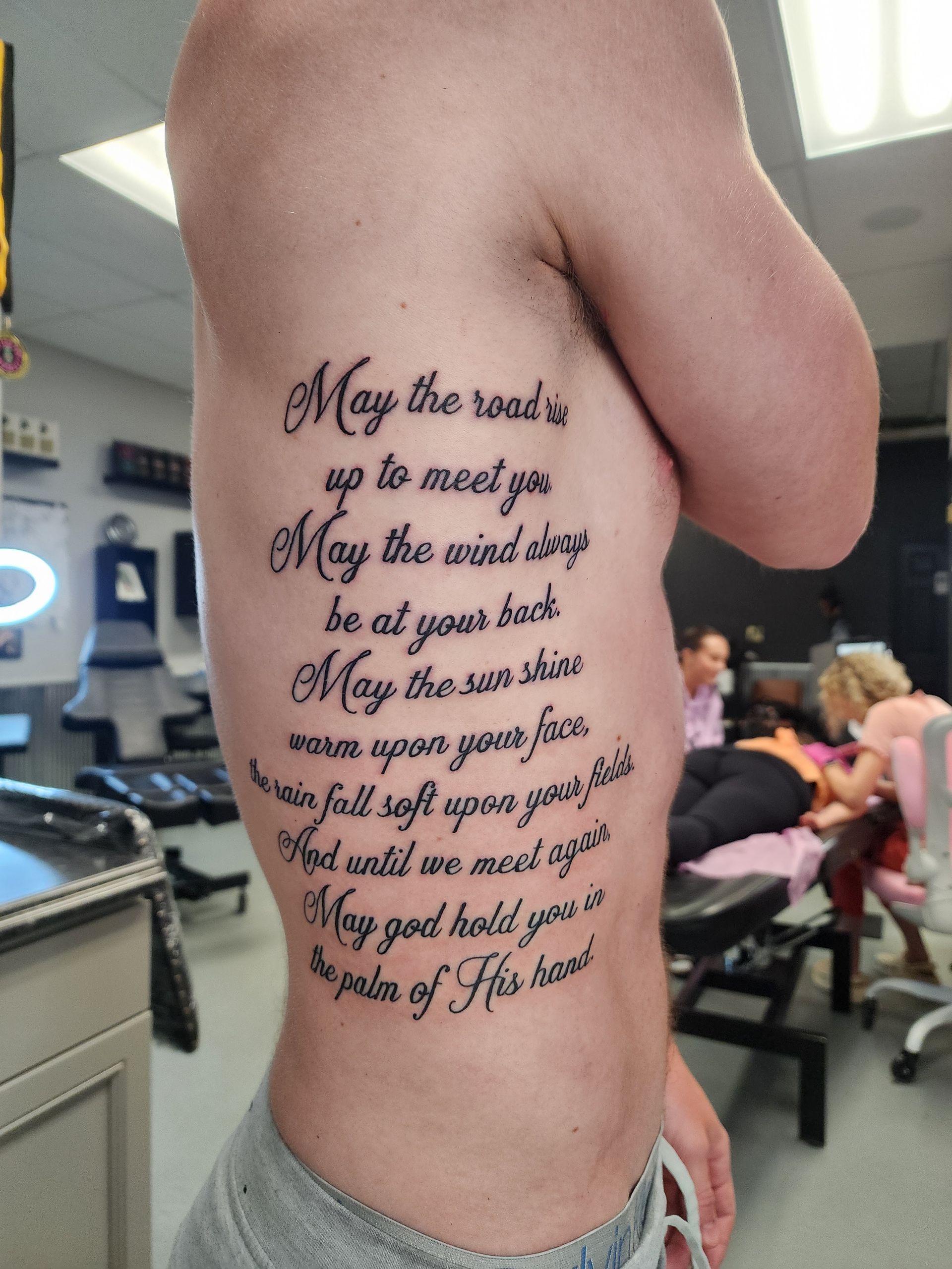 Tattoo on a person's side with a poem in cursive script. Background shows a tattoo parlor with people.