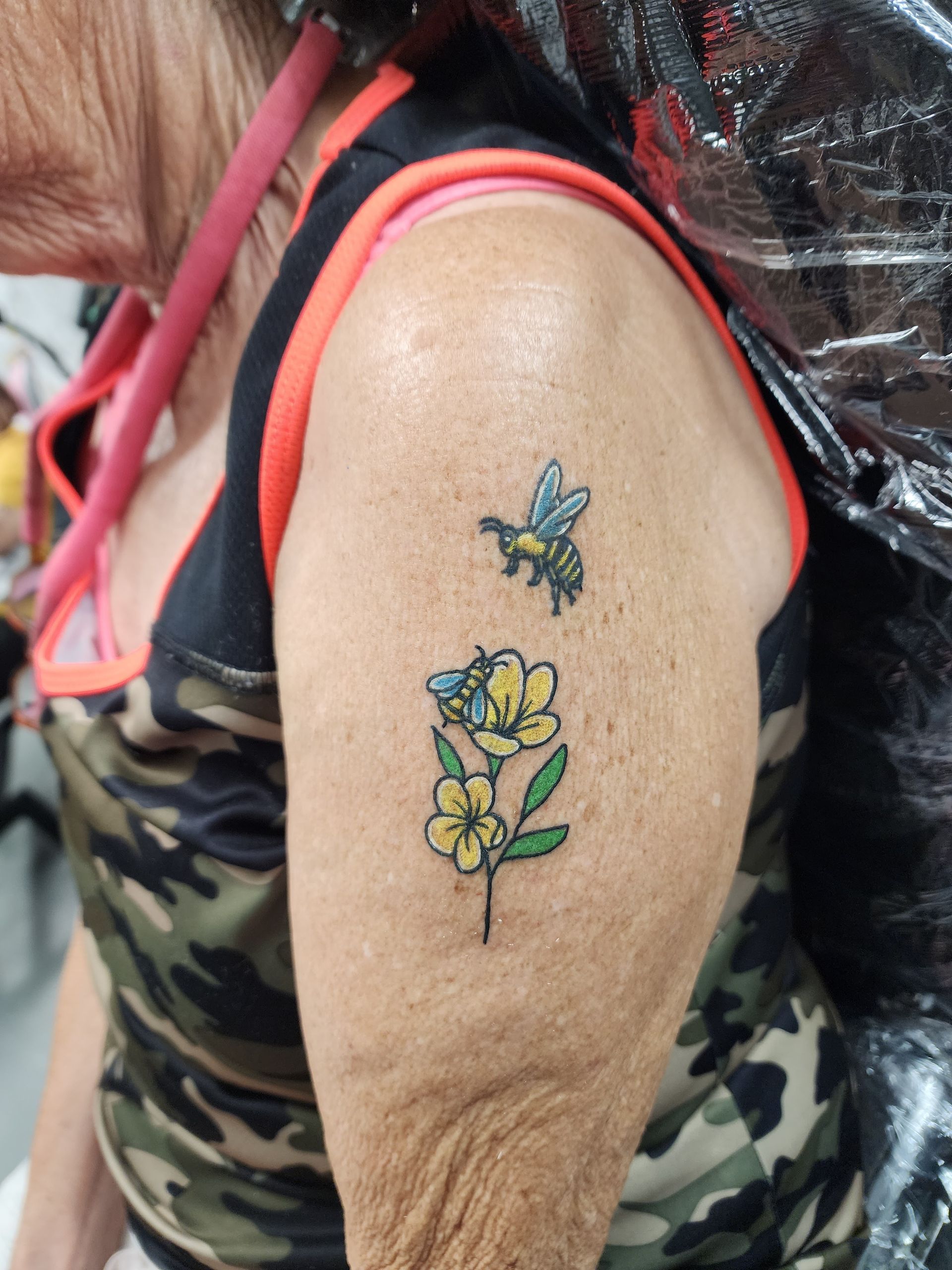 Tattoo of a bee and yellow flowers on a person's upper arm, with a camouflage tank top.